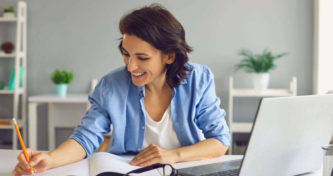 Uśmiechnięta kobieta w niebieskiej koszuli siedzi przy biurku, notuje coś w zeszycie i pracuje przy laptopie – przygotowuje biznesplan dla swojej firmy.