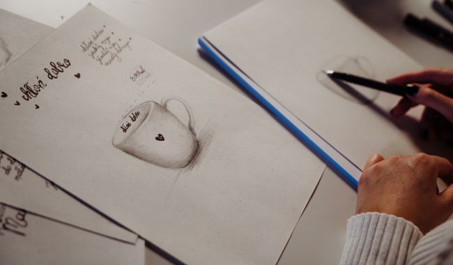 projekt kubków nadzwyczajnie.pl Proces projektowania kubków w pracowni marki Nadzwyczajnie.pl - zbliżenie na szkic kubka z sercem, ręce projektantki z ołówkiem oraz zapiski koncepcyjne na białym papierze