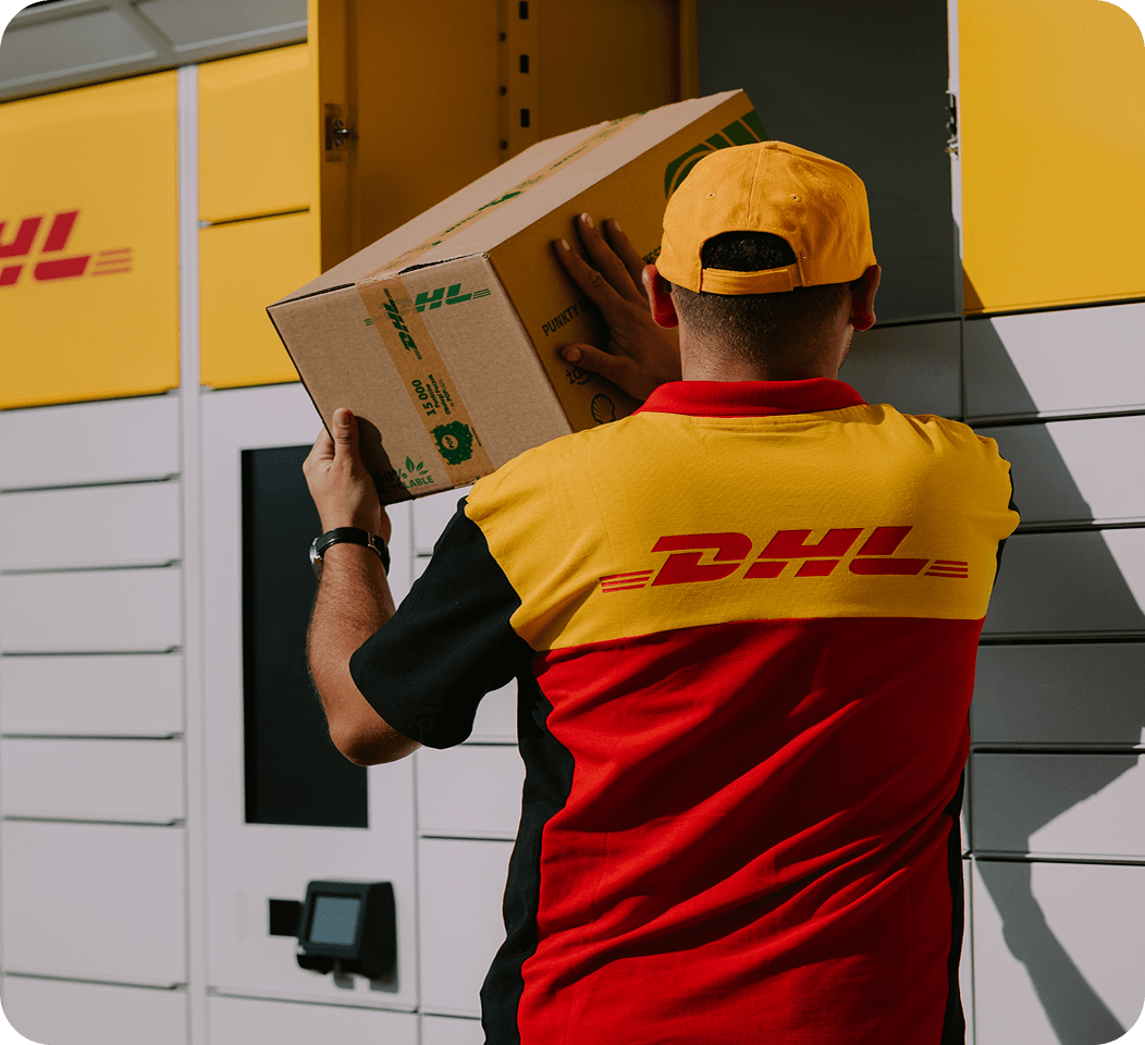 Kurier DHL w żółto-czerwonym stroju firmowym i czapce z daszkiem wkłada kartonową paczkę do otwartej skrytki żółto-białego automatu paczkowego DHL BOX 24/7.